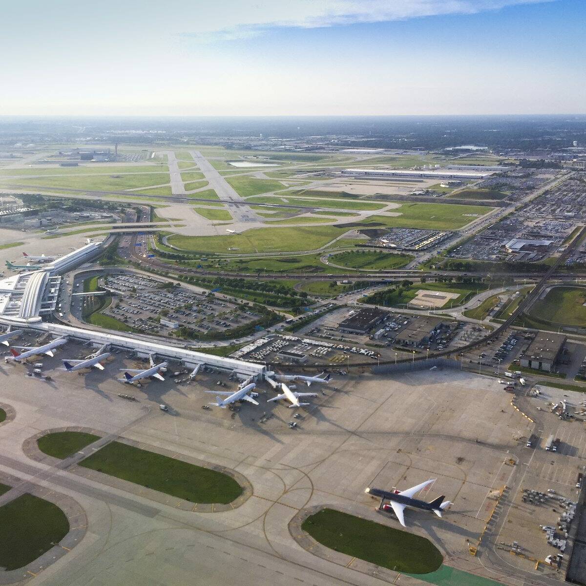 Aerial view of a busy airport with multiple airplanes parked at several terminals. The airport is surrounded by runways, taxiways, and various airport facilities. Beyond the airport, there is an expansive landscape with roads, buildings, and open areas.