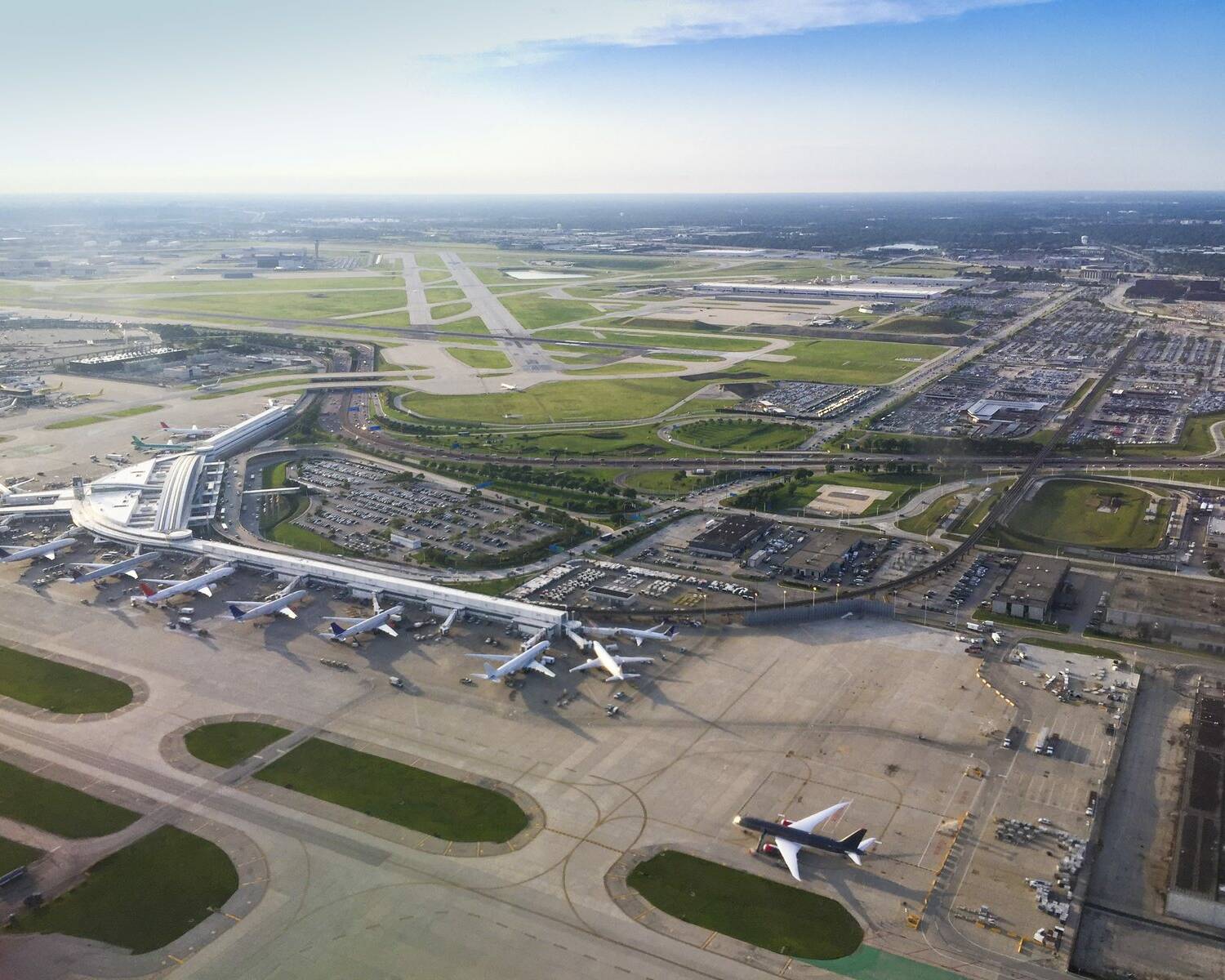 Aerial view of a busy airport with multiple airplanes parked at several terminals. The airport is surrounded by runways, taxiways, and various airport facilities. Beyond the airport, there is an expansive landscape with roads, buildings, and open areas.