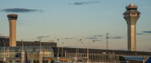 A panoramic view of an airport with two control towers standing tall against a clear sky. Terminal buildings with glass facades and boarding gates are visible below.