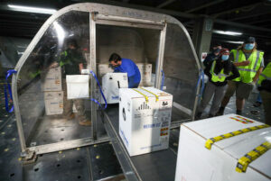 Workers load boxes labeled "medium" and featuring various vaccine-related symbols into a cargo hold. They wear safety vests and face masks during what seems to be an industrial operation, perhaps at an airport or warehouse. The activity reflects efforts mentioned in the President's Letter from December 2020.