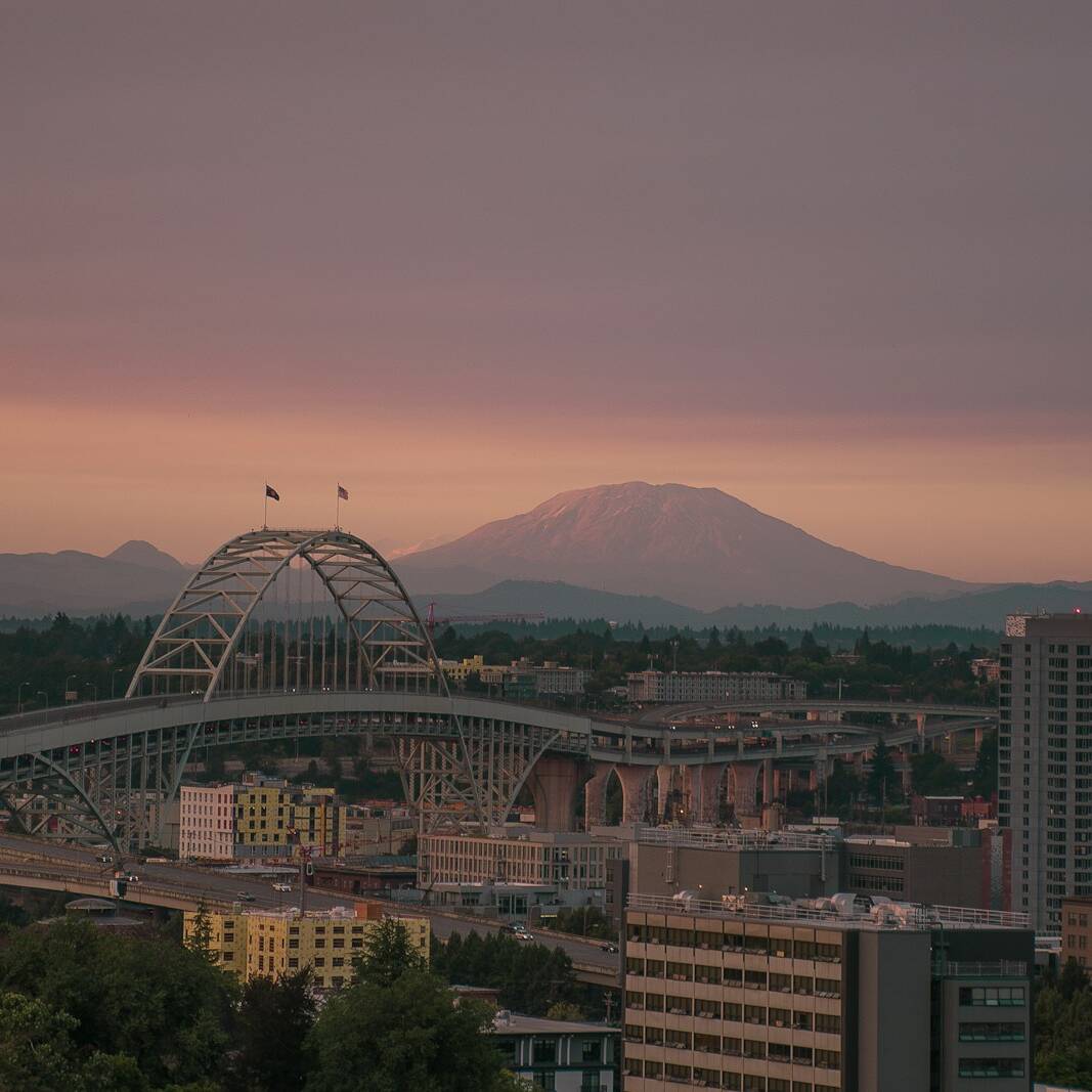 A scenic view of a city at dusk, featuring a prominent arch bridge spanning over a river with the backdrop of a vast, tree-covered landscape and a distant, towering mountain under a pastel-colored sky. High-rise buildings are visible near the bridge.