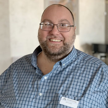 A man with glasses, a beard, and a plaid shirt smiles at the camera. He wears a name tag with indistinguishable text. The background is softly blurred, suggesting an indoor setting.