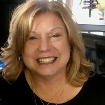 A woman with shoulder-length blonde hair smiles broadly. She is wearing a black top and a necklace. The background is slightly blurred, showing indoor elements like a window and chair.