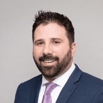 A man with short dark hair, a beard, and a mustache is smiling. He is wearing a blue suit jacket, a white dress shirt, and a light purple tie. The background is plain and light-colored.