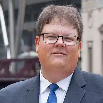 A man wearing black-framed glasses and a gray suit with a blue tie is smiling slightly. He has short, slightly tousled brown hair and stands in what appears to be an urban environment with modern and historic buildings in the background.