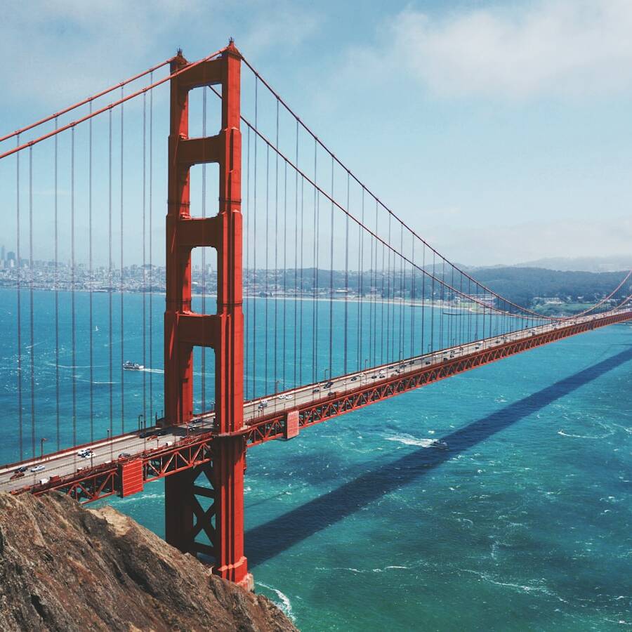 A wide-angle view of the Golden Gate Bridge with its iconic red-orange towers stretching across the blue waters of the San Francisco Bay. The city skyline is faintly visible in the background on a bright, clear day. A small boat is sailing beneath the bridge.