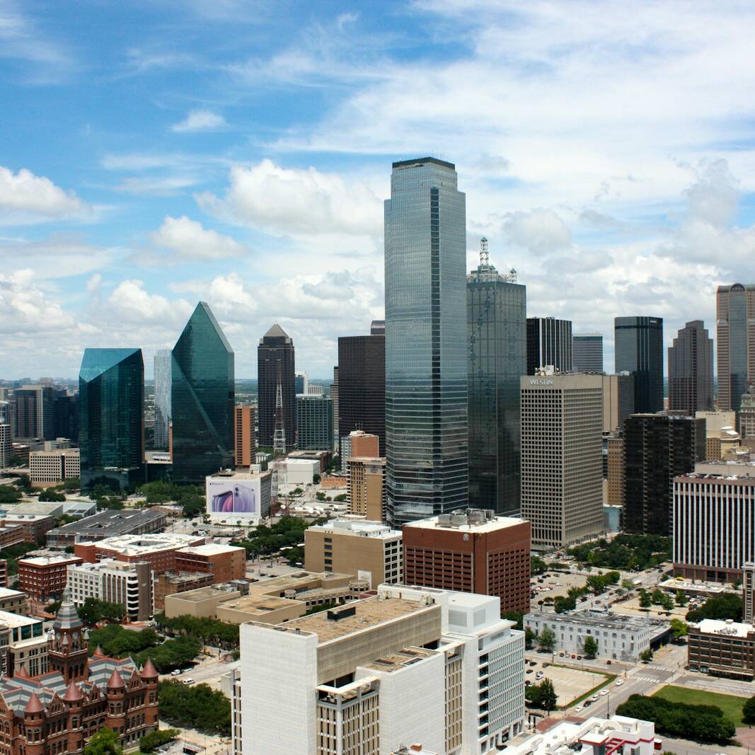 Aerial view of a city skyline with modern high-rise buildings under a partly cloudy sky. Notable structures include glass-paneled towers and a mix of older and newer architecture. The foreground shows a variety of shorter buildings and green spaces.
