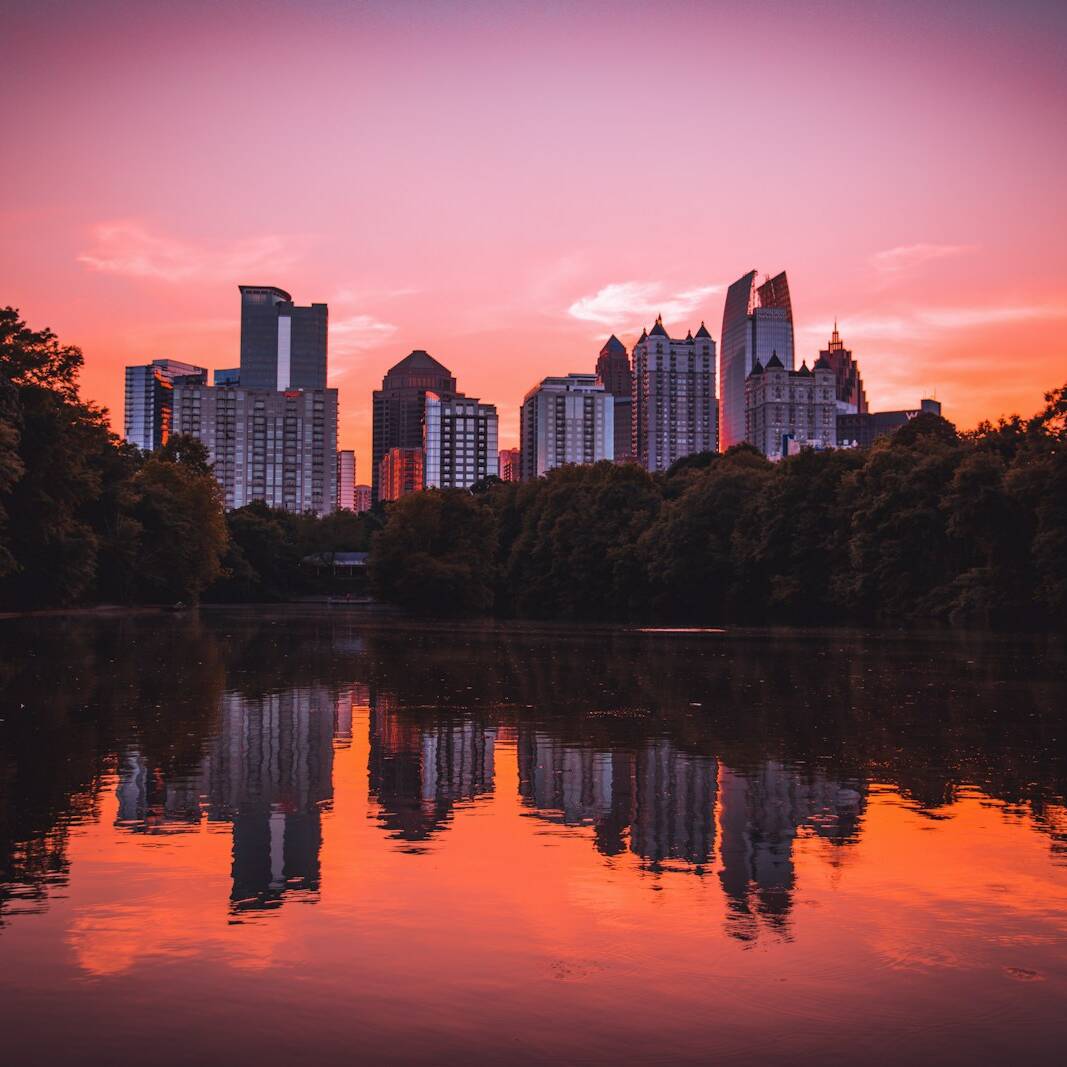 A city skyline with modern high-rise buildings is reflected in a calm body of water during a vibrant sunset. The sky is painted with shades of red, orange, and pink. Trees frame the water, creating a serene and picturesque scene.