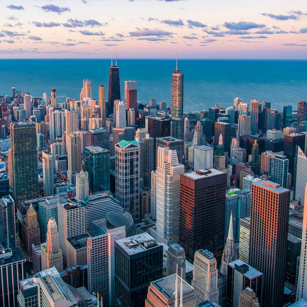 Aerial view of a vibrant cityscape during sunrise or sunset. Tall skyscrapers dominate the scene, reflecting the soft pink and blue hues of the sky. Lake Michigan is visible in the background, adding to the beautiful contrast of nature and urban architecture.