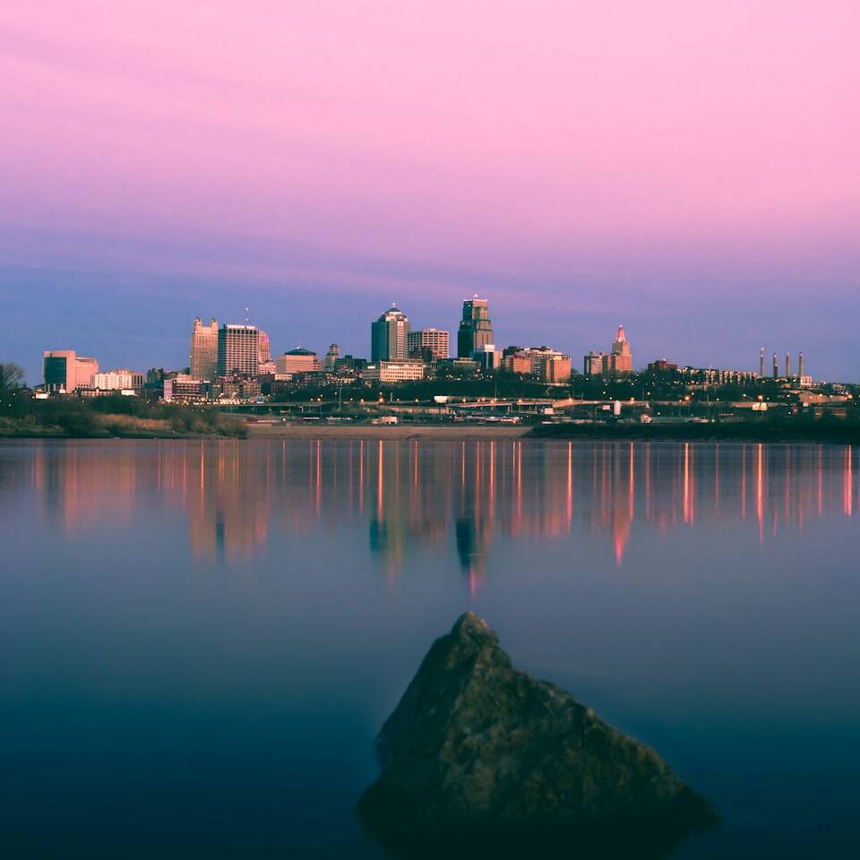 A serene cityscape at dusk with a clear, calm body of water in the foreground reflecting the twilight sky. The skyline features a variety of buildings, and the sky has a gradient of pink and purple hues. A rock protrudes from the water in the lower part of the image.