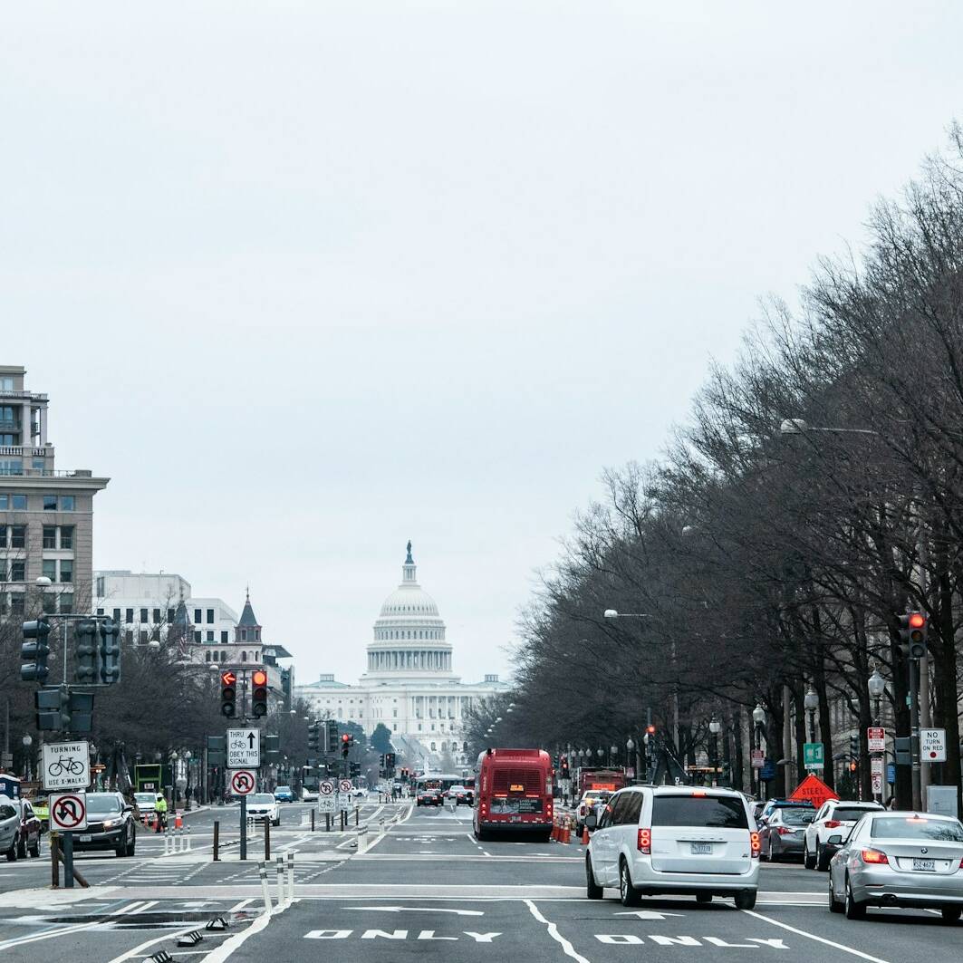A city street with several cars and a red bus, leading towards a domed building in the distance, which is the United States Capitol. Trees without leaves line both sides of the street, and buildings stand along the sidewalks. The sky is overcast.