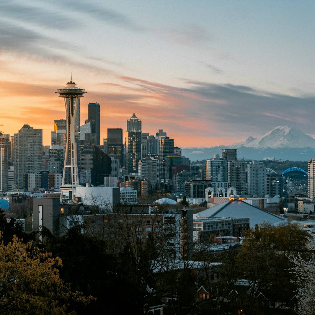 A scenic view of Seattle's skyline at sunset. The Space Needle stands prominently among the city buildings. In the background, Mount Rainier looms with snow-covered peaks under a partly cloudy sky. Trees and buildings in the foreground complete the urban landscape.