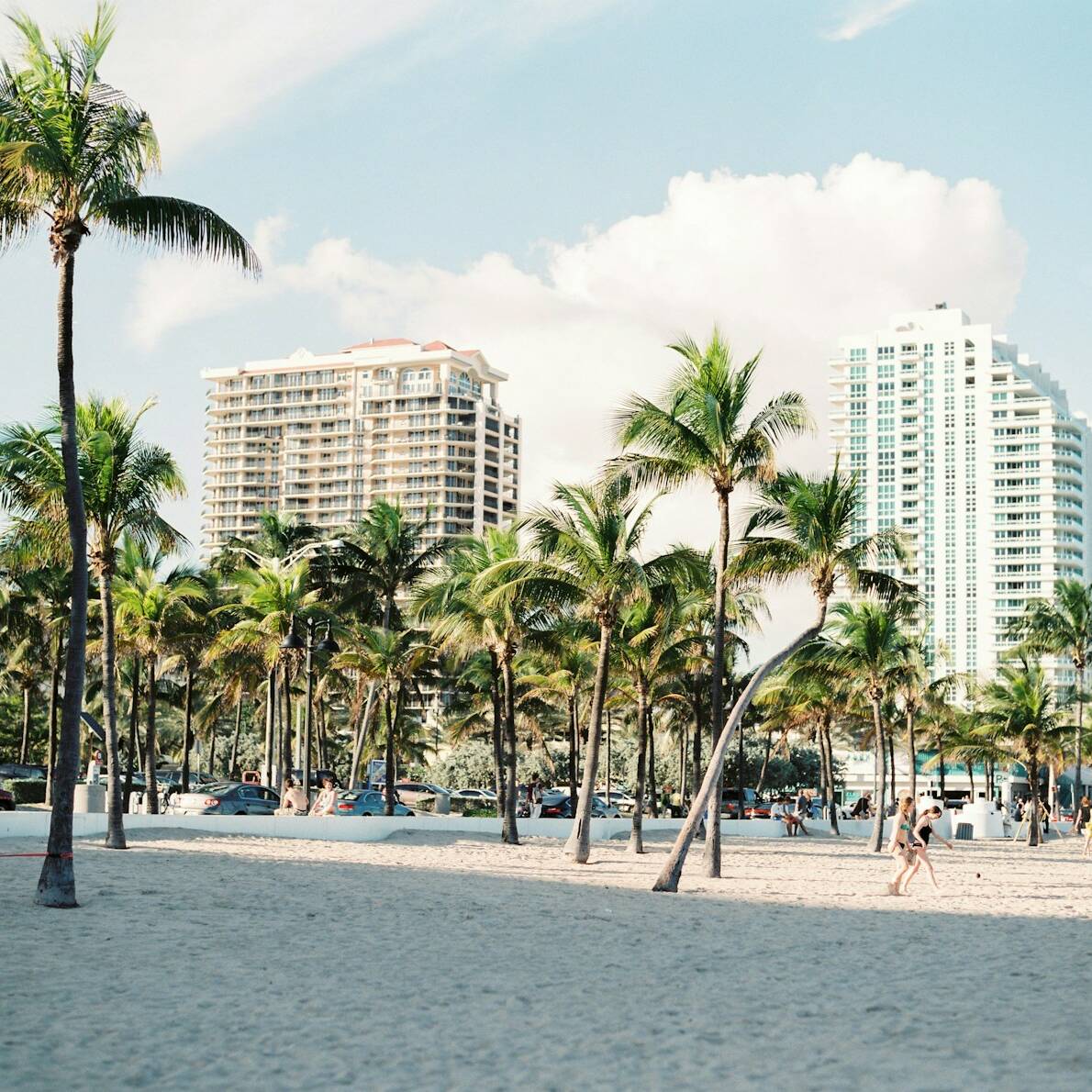 Sunny beachfront lined with tall palm trees and a row of high-rise buildings in the background. The sandy area has a few beachgoers strolling and cars parked near the edge. Blue skies with scattered clouds complete the tropical city landscape.