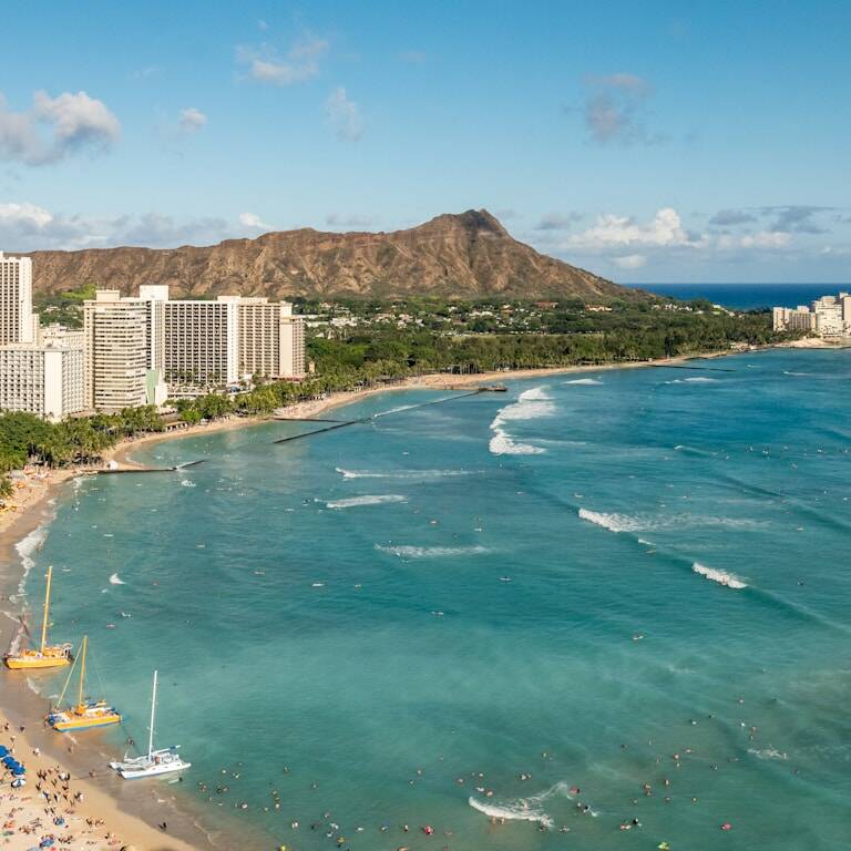Aerial view of Waikiki Beach in Honolulu, Hawaii, with numerous high-rise buildings along the shoreline. The turquoise ocean waves roll onto the sandy beach, which has patches of people and boats. Diamond Head crater is visible in the background under a blue sky.