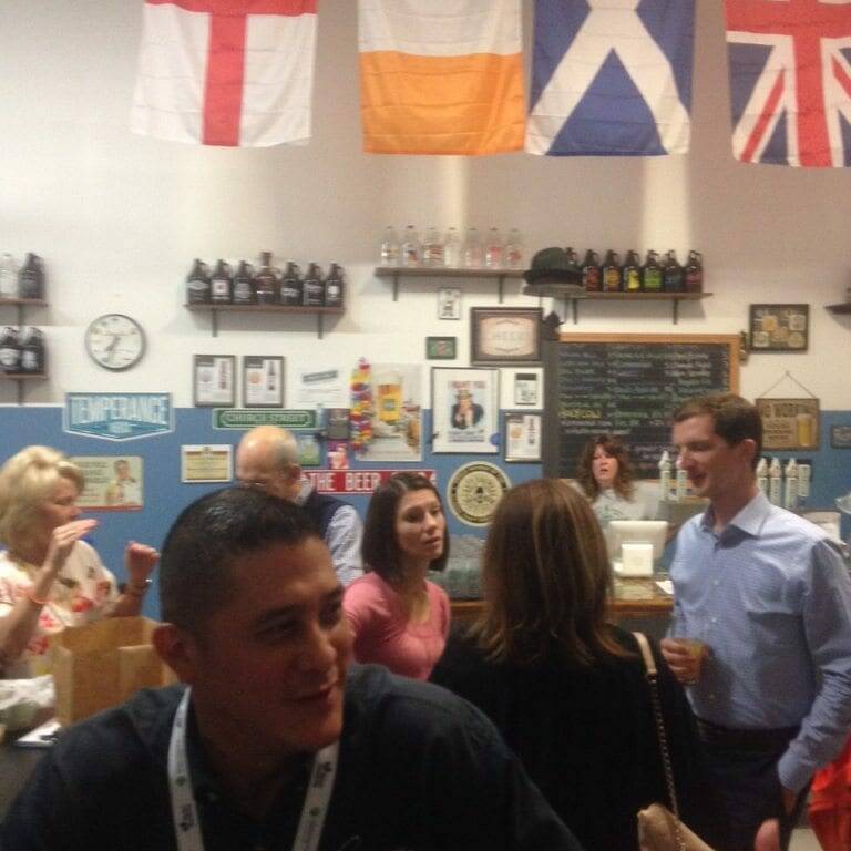 A diverse group of people socializing at a bar decorated with various international flags, photographs, and memorabilia on the walls. It's November 2022, and the spirit of Oktoberfest lingers as a bartender stands behind the counter, with some patrons holding drinks and conversing with one another.