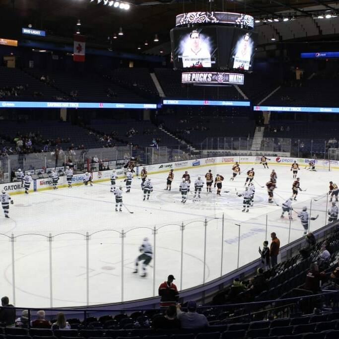 A hockey rink with players from the Chicago Wolves and another team warming up. Some players are skating, others are shooting pucks, while coaches are on ice. The stands have spectators scattered, and scoreboard screens display team logos and game information for the 2018 March Networking Night event.