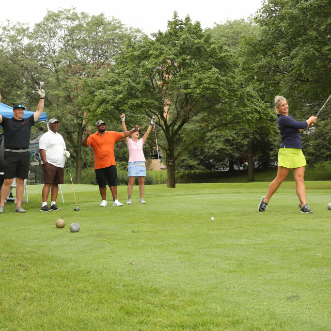 A woman swings a golf club on a lush green course during the 2021 IACAC Golf Outing while four individuals in the background cheer with their arms raised. A blue canopy and golf bags are also visible, surrounded by trees. The scene is lively and celebratory.