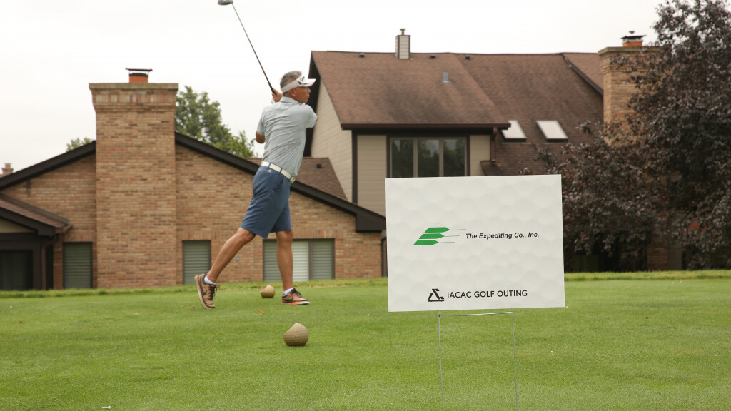 A golfer is captured mid-swing on a green. He is wearing a light-colored shirt, shorts, and a hat. In the foreground, there is a sign that reads “The Expediting Co., Inc. IACAC Golf Outing.” Brick buildings and trees are visible in the background.