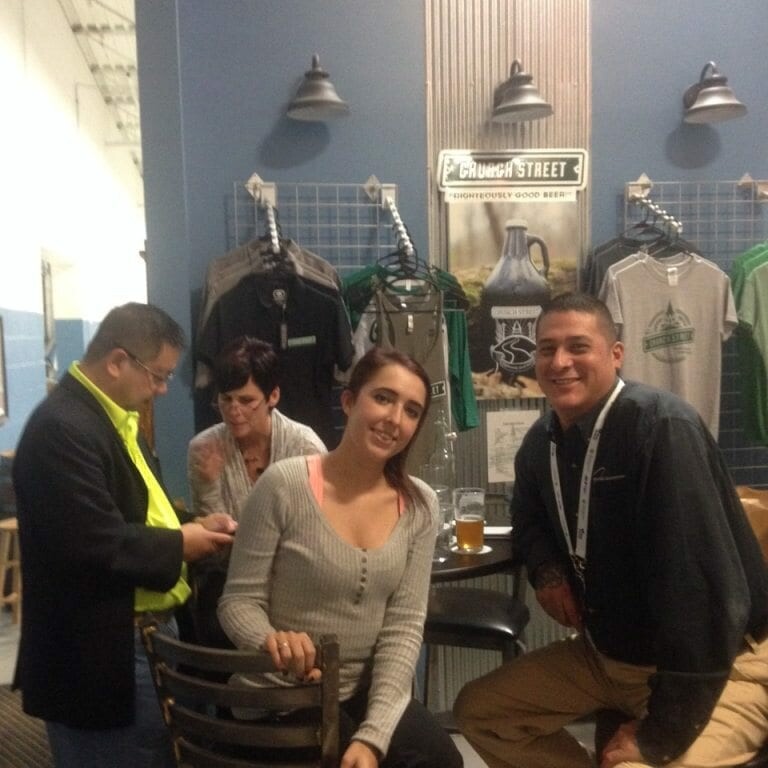 A group of four people are gathered in a casual indoor setting reminiscent of 2022 Oktoberfest. Two men and two women are present, with one woman sitting and smiling at the camera while the others engage in conversation or activities. The background features merchandise and festive decor.