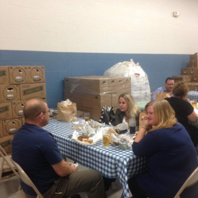 A group of four adults sit around a table covered in a blue and white checkered tablecloth, reminiscent of 2022 Oktoberfest, eating and chatting. The room has a plain interior with stacks of cardboard boxes and large wrapped bundles against the wall in the background.