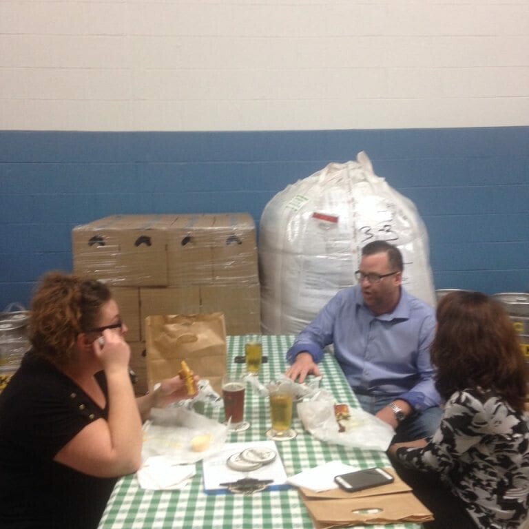 Three people converse while sitting at a table covered with a green and white checkered tablecloth, eating and drinking. The setting appears to be an industrial space, reminiscent of the 2022 Oktoberfest, with large sacks and boxes stacked in the background.