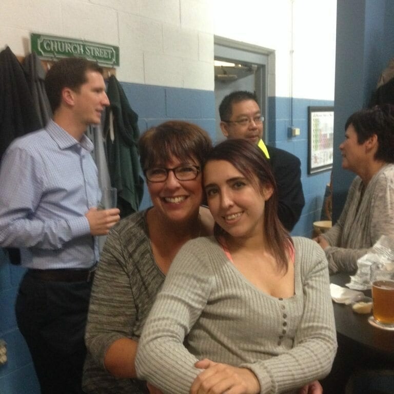 Two women are posing close together, smiling at the camera while sitting inside a room with blue and white walls. Behind them, three people are engaged in conversation, and a table with food and drinks is visible. A "Church Street" sign is on the wall in the background, capturing the festive spirit of November's Oktoberfest 2022.