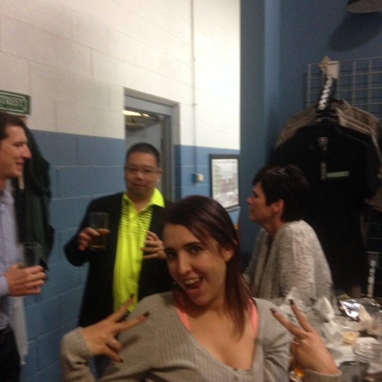 A woman in the foreground poses playfully with her hands raised in front of her chest. Behind her, three people are standing and talking, two of whom are holding drinks. The scene, reminiscent of a casual November 2022 gathering, takes place in a room with white and blue walls.