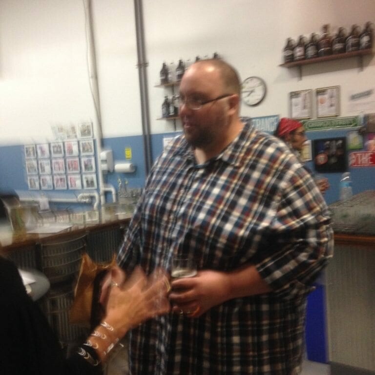 A man in a plaid shirt holding a drink stands indoors, conversing with someone whose back is to the camera. Shelves with bottles and photos are visible in the background. The scene appears to be in a casual setting like an Oktoberfest-themed bar or brewery.