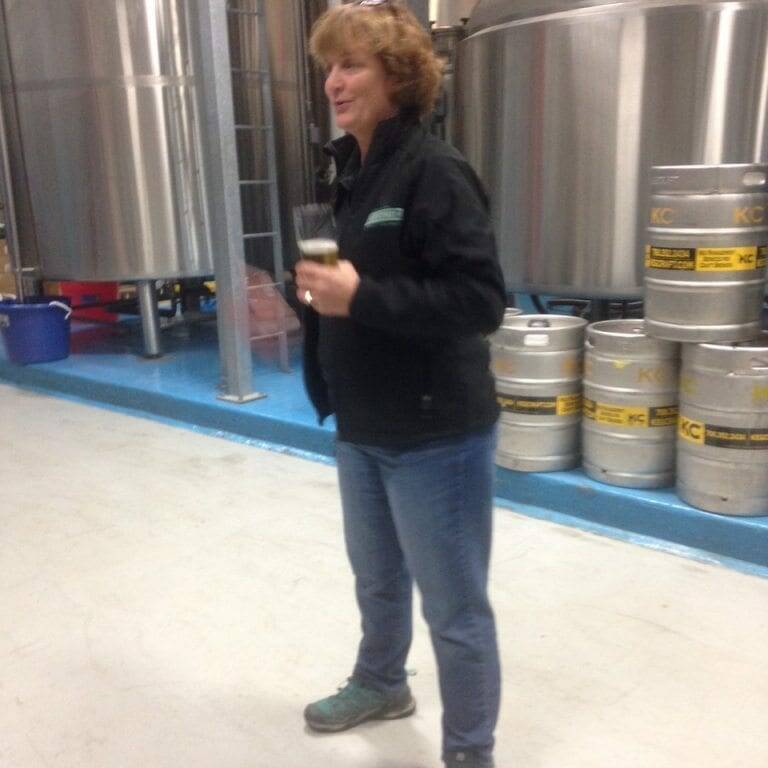 A person with short brown hair is standing on a brewery floor, holding a glass of beer, reminiscent of the festive spirit in 2022 Oktoberfest. They are wearing a black jacket, blue jeans, and sneakers. Large metal brewing tanks and several kegs create an industrial backdrop.