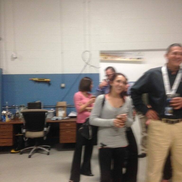 A group of people are standing in a room. Some are chatting, and one woman is holding a cup. The room has office furniture and a blue and white color scheme reminiscent of Oktoberfest. In the background, a woman is looking at her phone, and there are desks and chairs against the walls, setting an ambiance for November 2022.