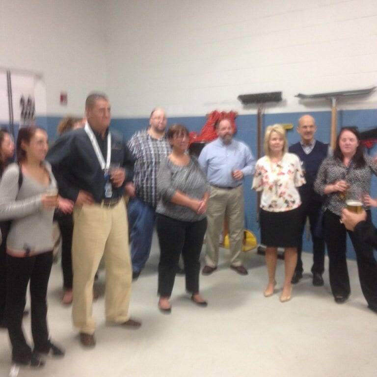 A group of people stands in a room with light-colored walls and cleaning equipment in the background. Some are holding drinks, possibly celebrating Oktoberfest, and appear to be engaged in casual conversation or listening to someone.