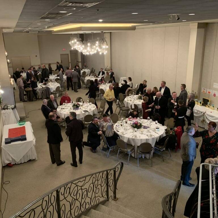 A formal banquet hall filled with people standing and sitting around large round tables adorned with white tablecloths. A staircase with ornate railings is visible in the foreground, hinting at a holiday luncheon atmosphere. Chandeliers hang from the ceiling, creating a sophisticated ambiance.