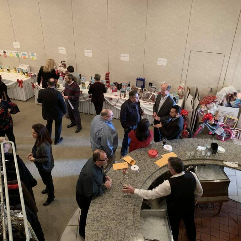 At the 2018 Holiday Luncheon, a group of people are gathered in a room for the event. They are browsing various tables displaying items, possibly for a raffle or auction. In the foreground, a bartender is serving drinks behind a curved counter. The atmosphere is lively and social.