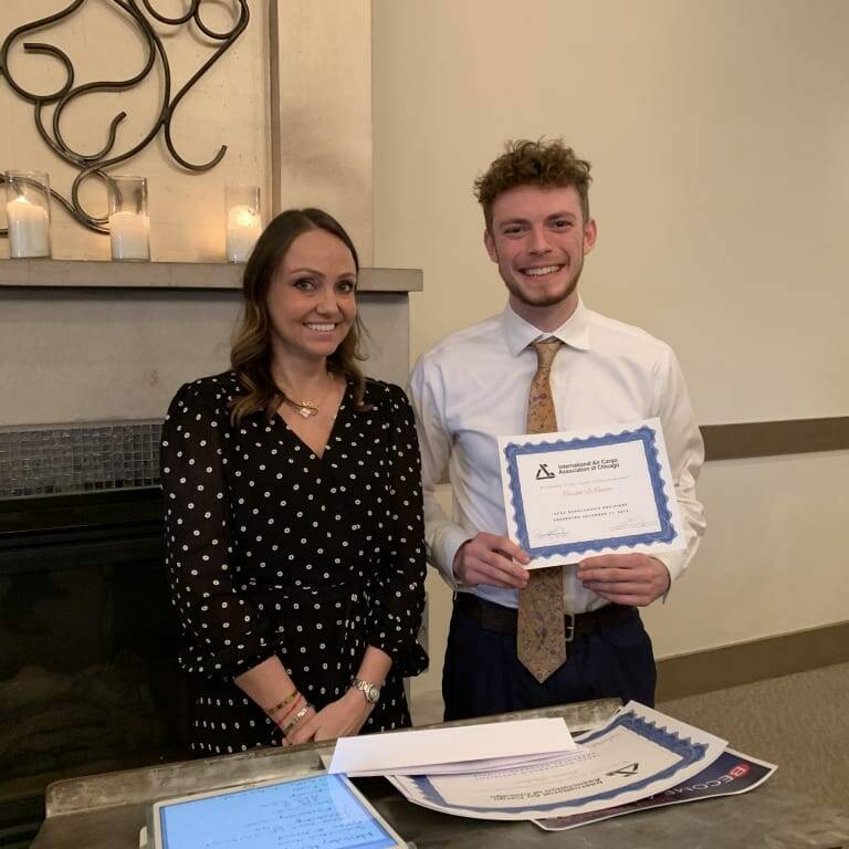 A man in a white shirt and tie, holding a certificate and smiling, stands next to a woman in a black dress with white polka dots. They are indoors, next to a fireplace with candles on the mantel. A table in front of them holds papers and certificates from the 2018 holiday luncheon.