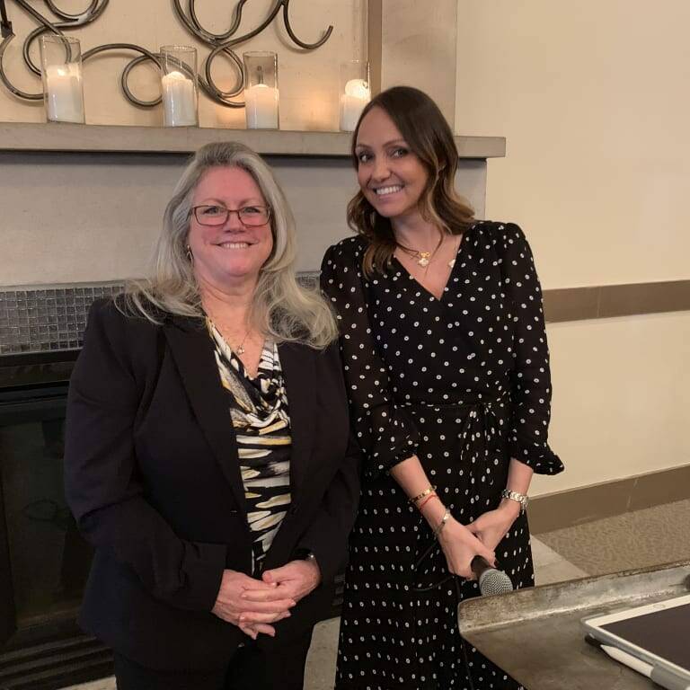 Two women are standing indoors in front of a fireplace with candles on the mantle. The woman on the left has long, gray hair and is wearing a black blazer, while the woman on the right, dressed in a black dress with white polka dots and holding a microphone, is speaking at the 2018 holiday luncheon.