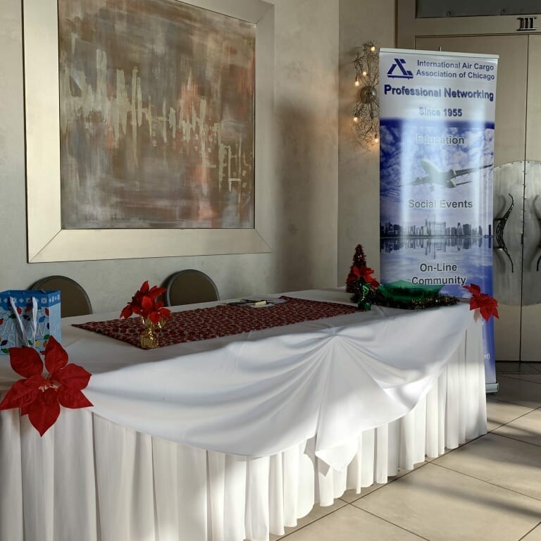 A booth featuring a white tablecloth with red accents, adorned with poinsettias. Behind the table is a roll-up banner promoting the International Air Cargo Association of Chicago's 2018 Holiday Luncheon, highlighting networking, education, social events, and an online community.