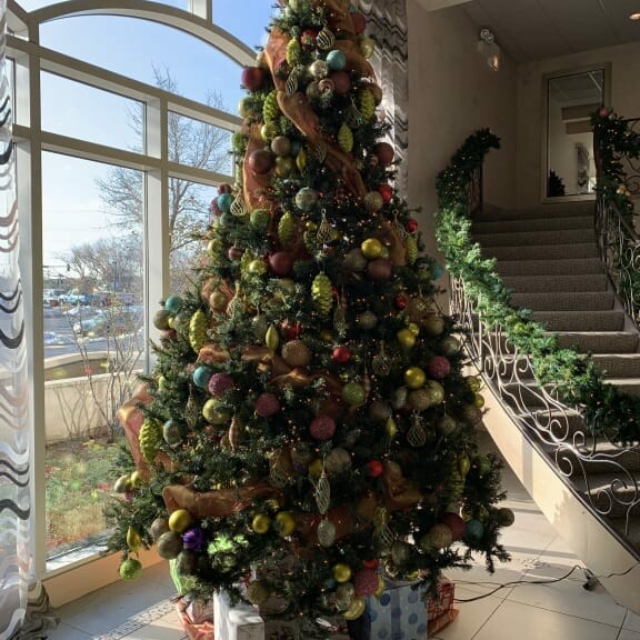 A decorated Christmas tree stands in the corner of a room with large, arched windows. The tree is adorned with various ornaments, ribbons, and lights. Wrapped presents are placed at its base. A staircase with a garland railing is visible to the right, set for a festive holiday luncheon in 2018.