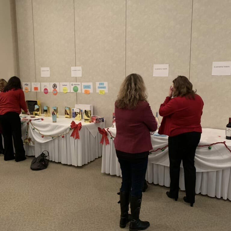 People are viewing items arranged on two tables draped in white tablecloths decorated with holiday-themed accents. Signs and small gifts are displayed on the tables, reminiscent of a festive luncheon, and the individuals are casually dressed, engaging with the items.