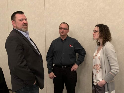 Three people are standing and conversing in what appears to be a professional setting. The man on the left is wearing a dark suit, the man in the middle is in a dark shirt with a logo, and the woman on the right is in a light cardigan and floral blouse, all possibly discussing plans for an upcoming holiday luncheon.