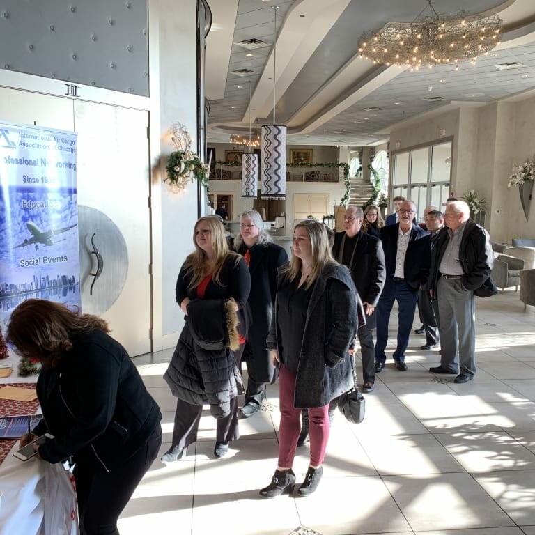 A group of people in winter attire stand in line inside a well-lit and decorated venue. They appear to be checking in at a reception table covered with informational materials near a holiday-themed display. A banner in the background promotes the 2018 Holiday Luncheon event.