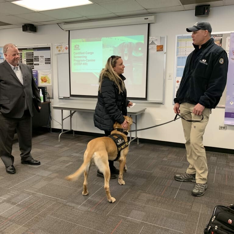 Three people are standing in an office. A woman is interacting with a service dog wearing a vest labeled "Certified Cargo Service." Two men are observing; one, from K2 Solutions, is holding the dog's leash. A presentation from the 2019 February Luncheon is displayed on a screen in the background.