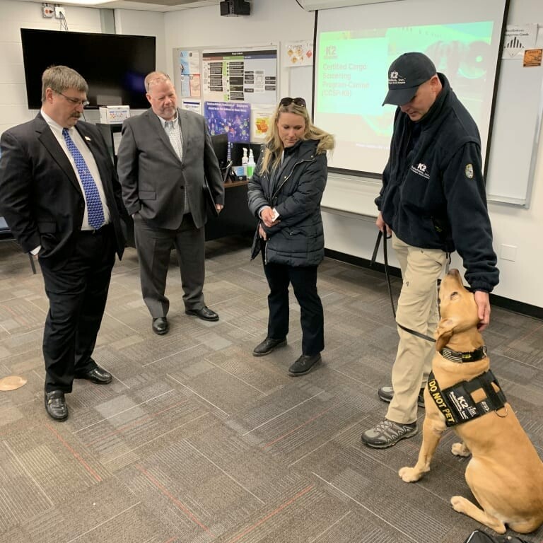 Four people stand in a room with one of them, wearing a black jacket and cap, holding a harnessed service dog sitting beside them. The group appears to be having a conversation at the 2019 February Luncheon event. In the background, a presentation slide is displayed on a screen.