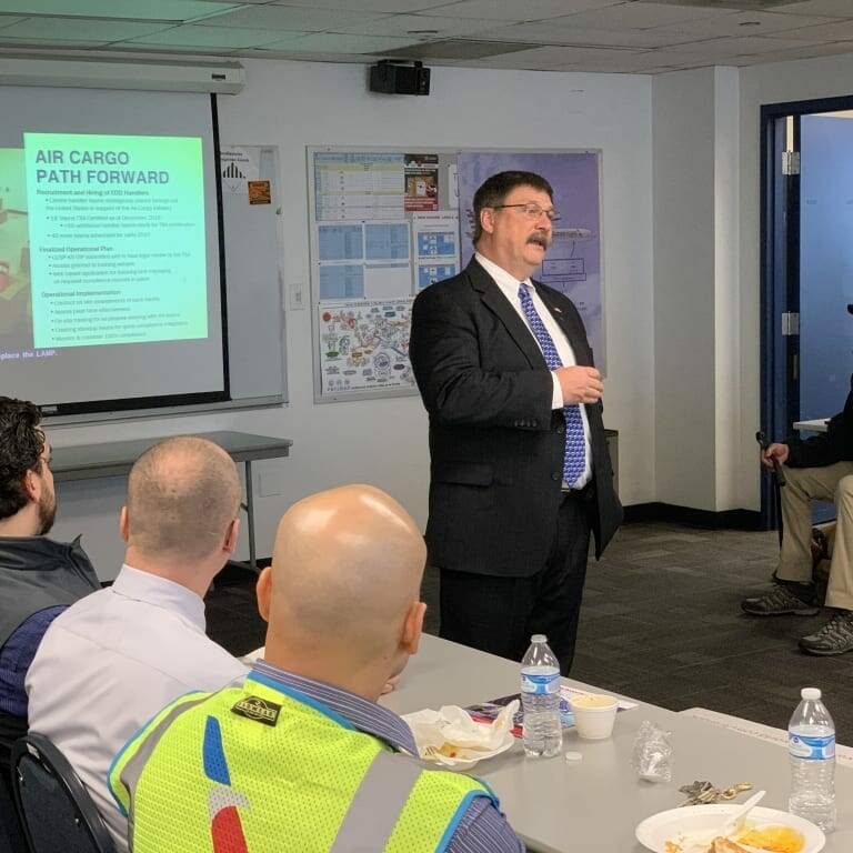 A man in a suit stands in front of an audience, giving a presentation on "Air Cargo Path Forward" at the 2019 February Luncheon by K2 Solutions. The room has several people sitting at desks with water bottles and food. A screen behind him displays presentation slides, with an exit sign visible in the background.