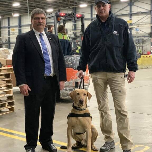 Two men stand in a warehouse next to a yellow Labrador wearing a K-9 harness. The man on the left is dressed in a suit and tie, while the man on the right wears a black jacket and beige pants, holding the dog’s leash. A forklift operates in the background during the 2019 February Luncheon by K2 Solutions.