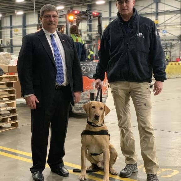 Two men stand in a warehouse with a yellow Labrador Retriever dog wearing a K9 harness. The man on the left is dressed in a suit, while the man on the right is in casual attire and baseball cap. A forklift and warehouse shelves are visible in the background, reminiscent of the 2019 February Luncheon event.