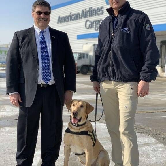 Two men stand beside a yellow Labrador wearing a service dog vest. The man on the left is in a business suit and tie, while the man on the right is in a casual jacket and cap. They are outside an American Airlines Cargo facility, likely after attending the February 2019 luncheon hosted by K2 Solutions.