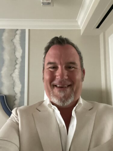 A middle-aged man with short gray hair and a neatly trimmed goatee smiles at the camera. William Gerst, a dedicated board member, wears a light-colored suit jacket and a white dress shirt. The background shows part of a room with a piece of abstract wall art and minimal decor.