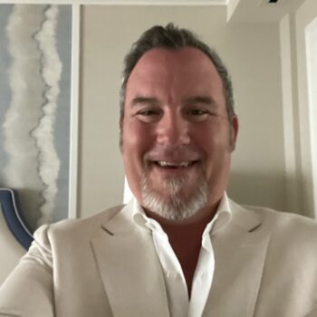 A middle-aged man with short gray hair and a neatly trimmed goatee smiles at the camera. William Gerst, a dedicated board member, wears a light-colored suit jacket and a white dress shirt. The background shows part of a room with a piece of abstract wall art and minimal decor.