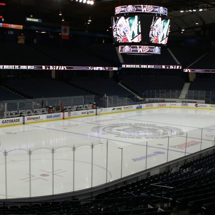 An empty indoor ice hockey arena with a clean, illuminated rink. Bright lights and a scoreboard hang from the ceiling above the rink. The seating areas appear dark, and various advertisements are visible along the boards surrounding the ice, including a banner for the Chicago Wolves' 2018 March Networking Night.