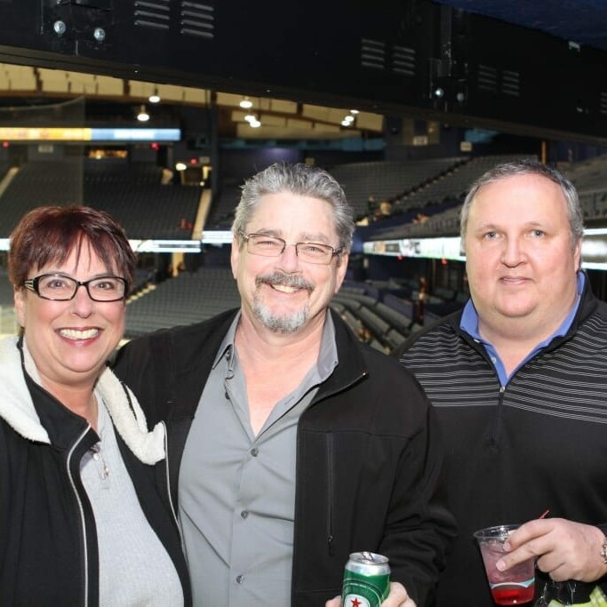 Three people are standing and smiling at the Chicago Wolves' Networking Night in an indoor stadium. From left to right: a woman wearing glasses, a man with glasses in a gray shirt, and a man in a black and gray shirt. The stadium seats are visible in the background. The man on the left holds a can, and the man on the right holds a cup.